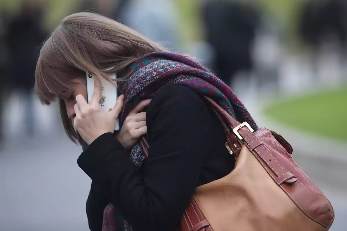 Una persona hablando por teléfono mientras camina al aire libre con un bolso marrón y una bufanda colorida.