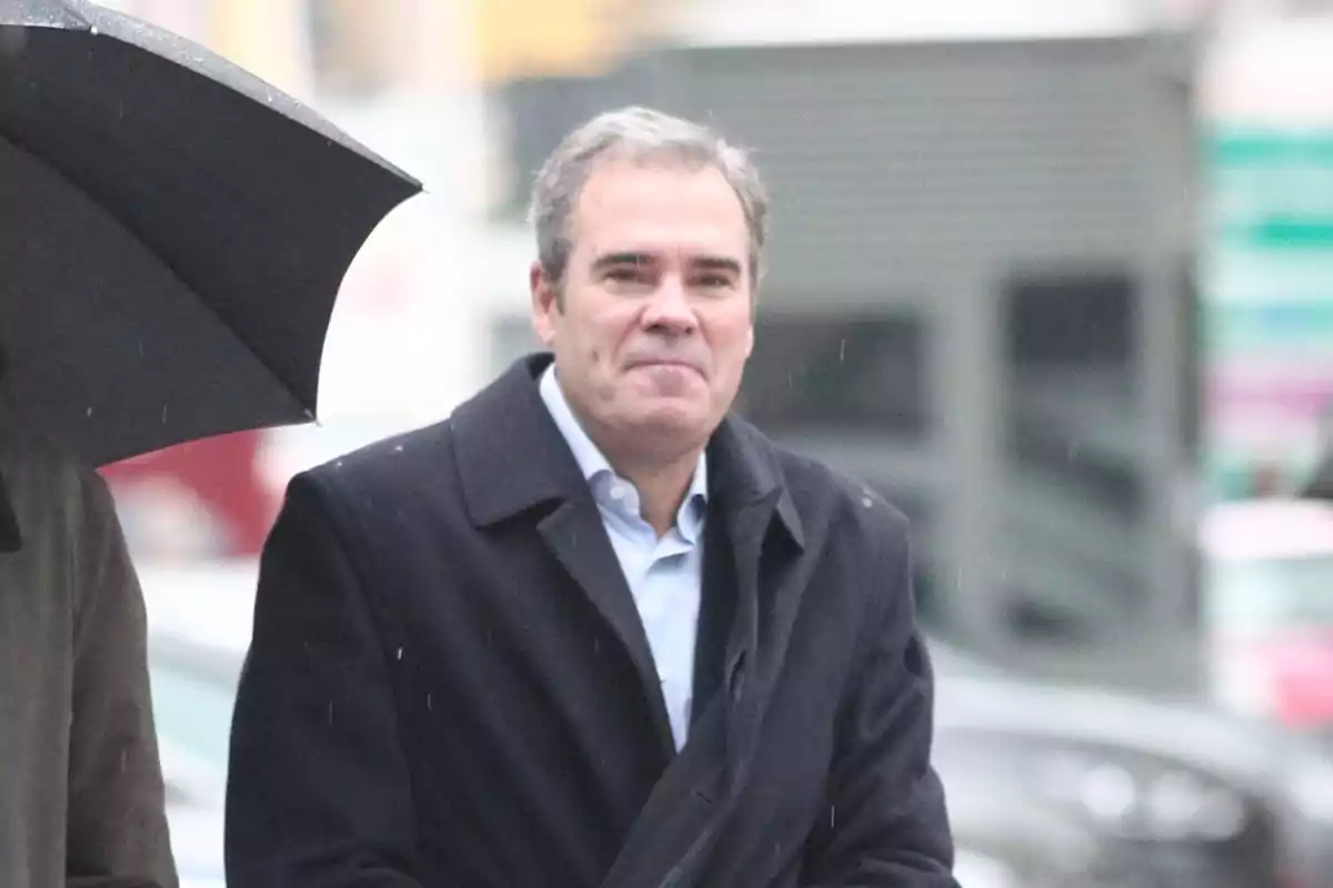 Hombre de cabello canoso con abrigo oscuro caminando bajo la lluvia junto a una persona con paraguas negro Hombre de cabello canoso con abrigo oscuro caminando bajo la lluvia junto a una persona con paraguas negro