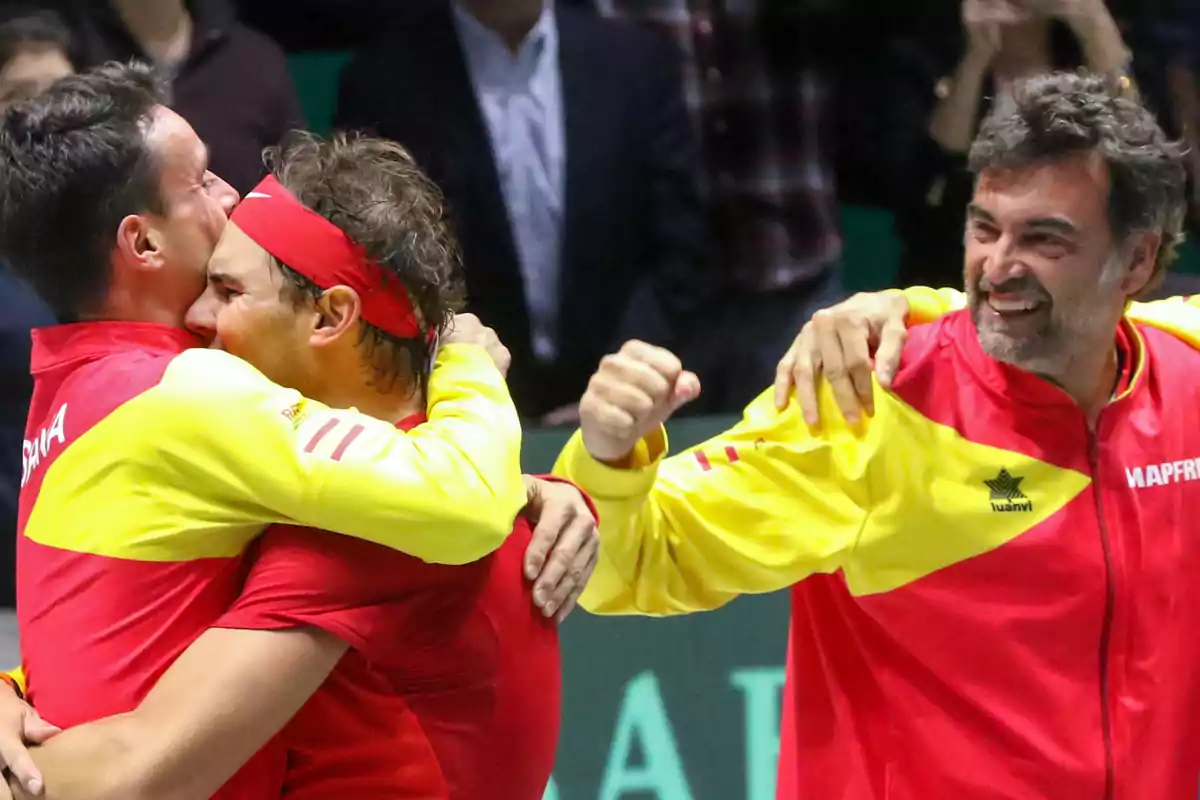 Tres personas con chaquetas rojas y amarillas celebran abrazándose y sonriendo en un evento deportivo.