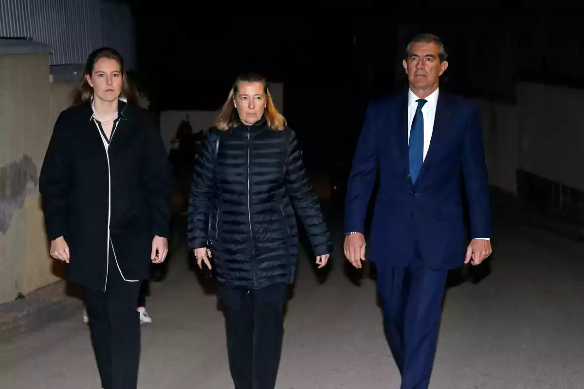 Tres personas caminando juntas por la noche, una mujer joven a la izquierda, una mujer de mediana edad en el centro y un hombre de traje a la derecha.