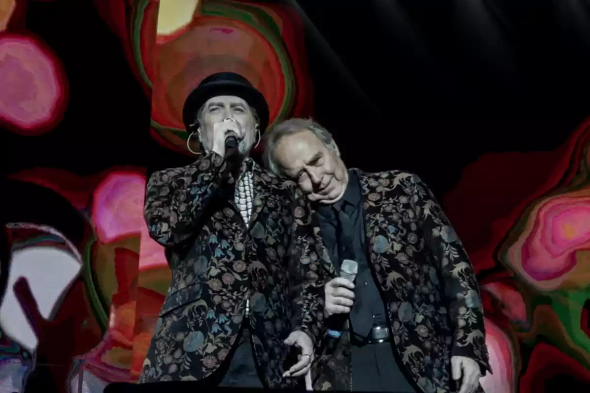 Dos personas en el escenario, una cantando con un micrófono y la otra apoyando su cabeza en el hombro de la primera, ambas con chaquetas estampadas y un fondo colorido. Dos personas en el escenario, una cantando con un micrófono y la otra apoyando su cabeza en el hombro de la primera, ambas con chaquetas estampadas y un fondo colorido.