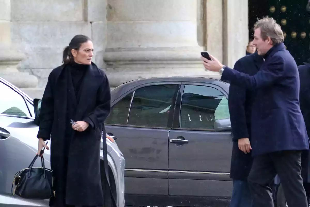 Una mujer vestida de negro sostiene un bolso mientras un hombre le toma una foto con su teléfono móvil frente a un edificio de piedra. Una mujer vestida de negro sostiene un bolso mientras un hombre le toma una foto con su teléfono móvil frente a un edificio de piedra.