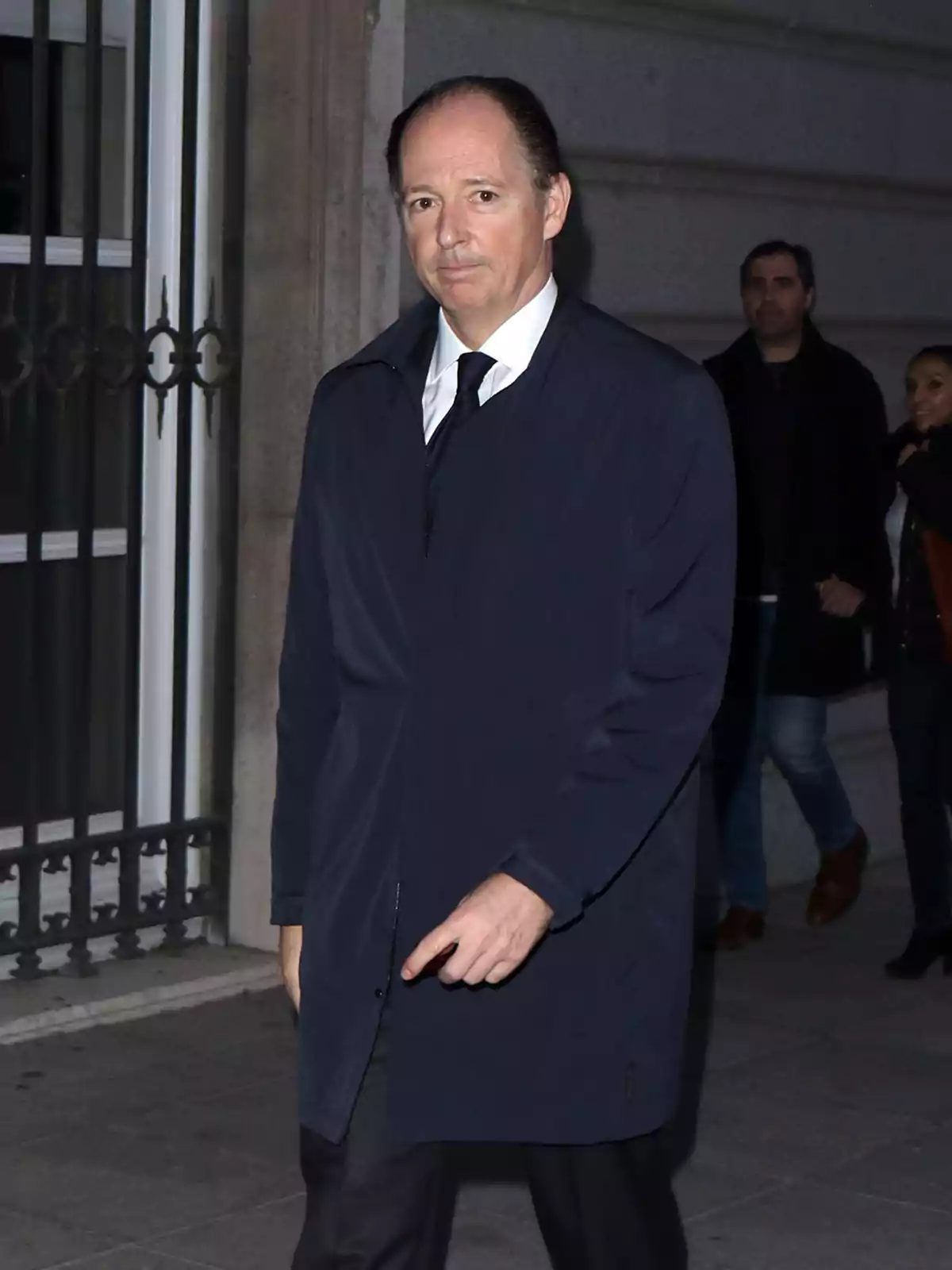 Hombre de mediana edad con abrigo azul oscuro y corbata caminando por la acera frente a un edificio de piedra Hombre de mediana edad con abrigo azul oscuro y corbata caminando por la acera frente a un edificio de piedra
