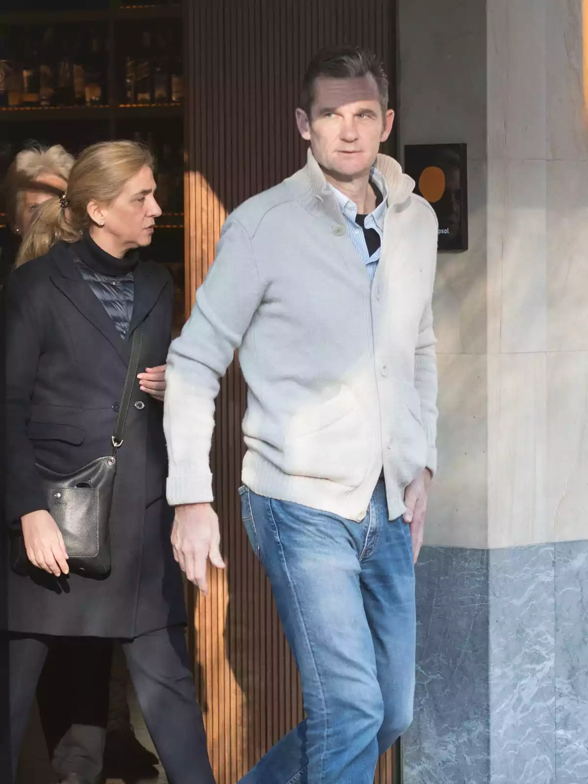 Un hombre con suéter claro y jeans camina junto a una mujer rubia con abrigo oscuro y bolso negro saliendo de un edificio Un hombre con suéter claro y jeans camina junto a una mujer rubia con abrigo oscuro y bolso negro saliendo de un edificio