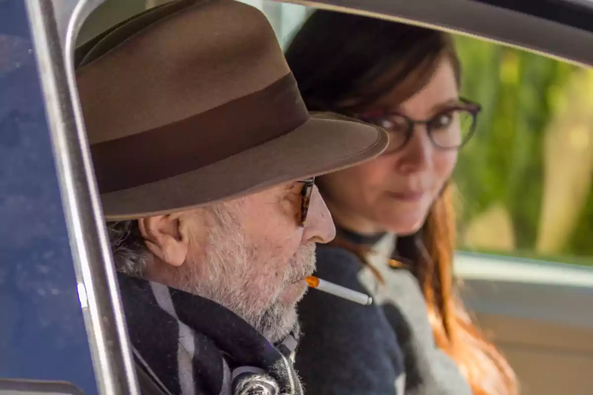 Hombre mayor con sombrero y gafas fumando un cigarro dentro de un coche junto a una mujer joven con gafas Hombre mayor con sombrero y gafas fumando un cigarro dentro de un coche junto a una mujer joven con gafas