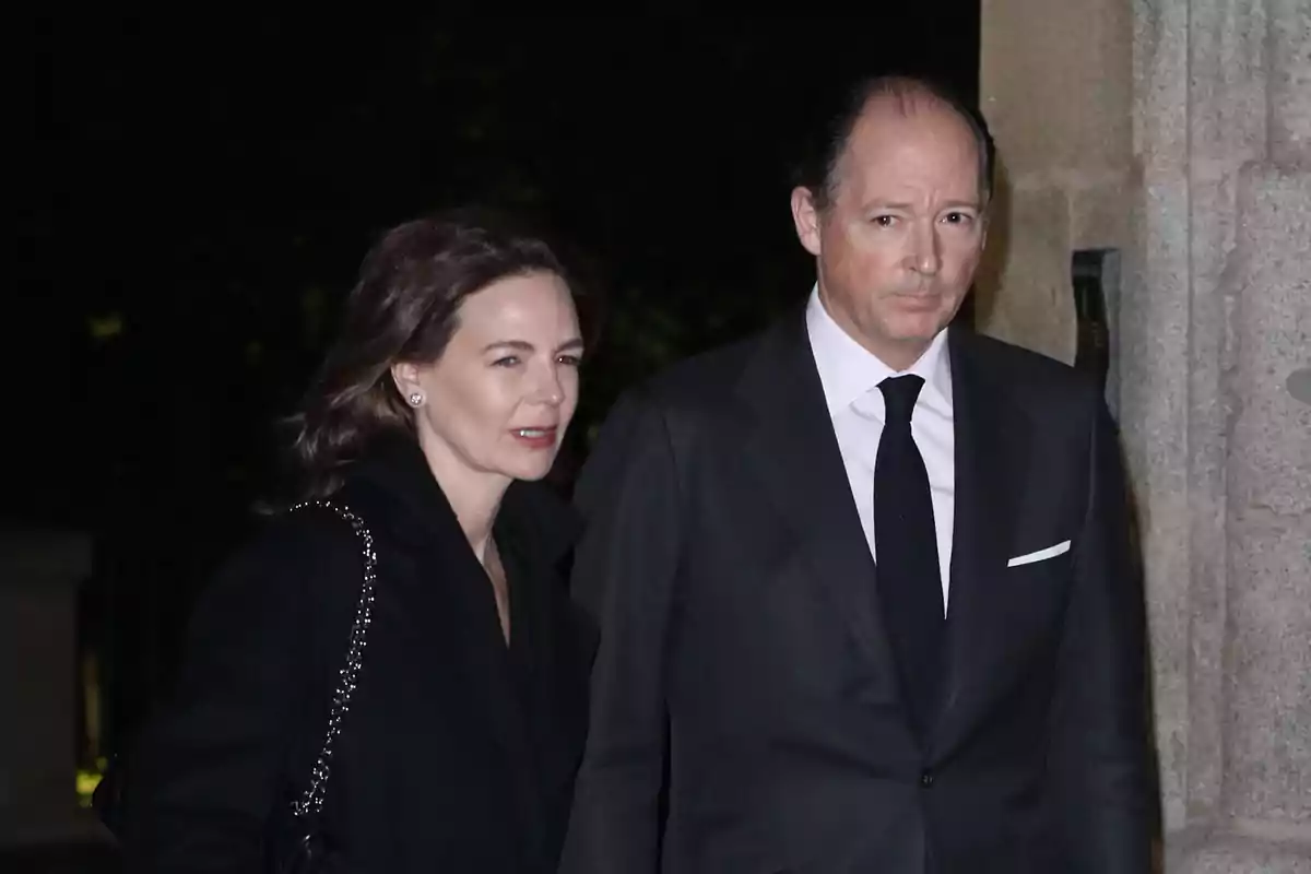 Una pareja elegantemente vestida camina junta en un entorno nocturno, ambos con expresión seria y ropa formal oscura. Una pareja elegantemente vestida camina junta en un entorno nocturno, ambos con expresión seria y ropa formal oscura.