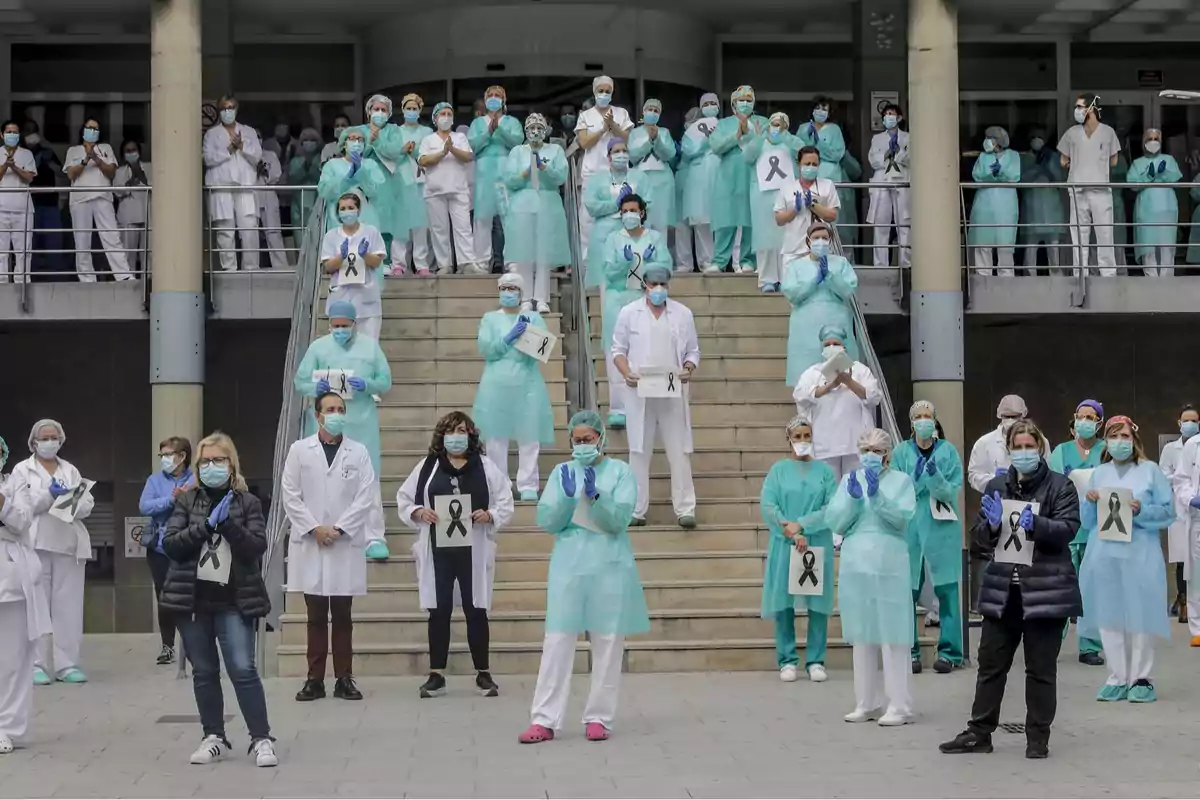 Un grupo de profesionales de la salud con mascarillas y batas se encuentra de pie en una escalera, algunos sostienen carteles con lazos negros.