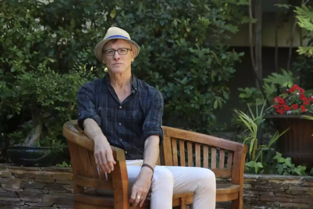 Hombre con sombrero y gafas sentado en un banco de madera en un jardín.