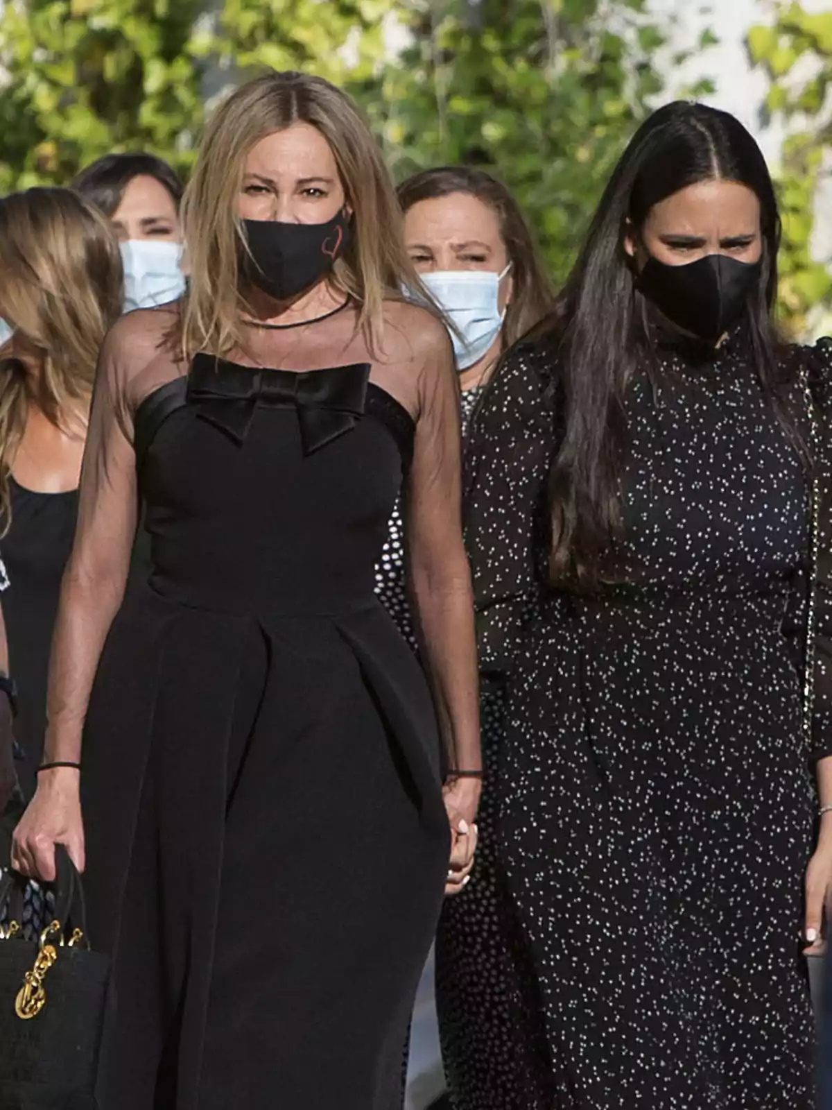 Un grupo de mujeres vestidas de negro, algunas con mascarillas, caminan al aire libre. Un grupo de mujeres vestidas de negro, algunas con mascarillas, caminan al aire libre.