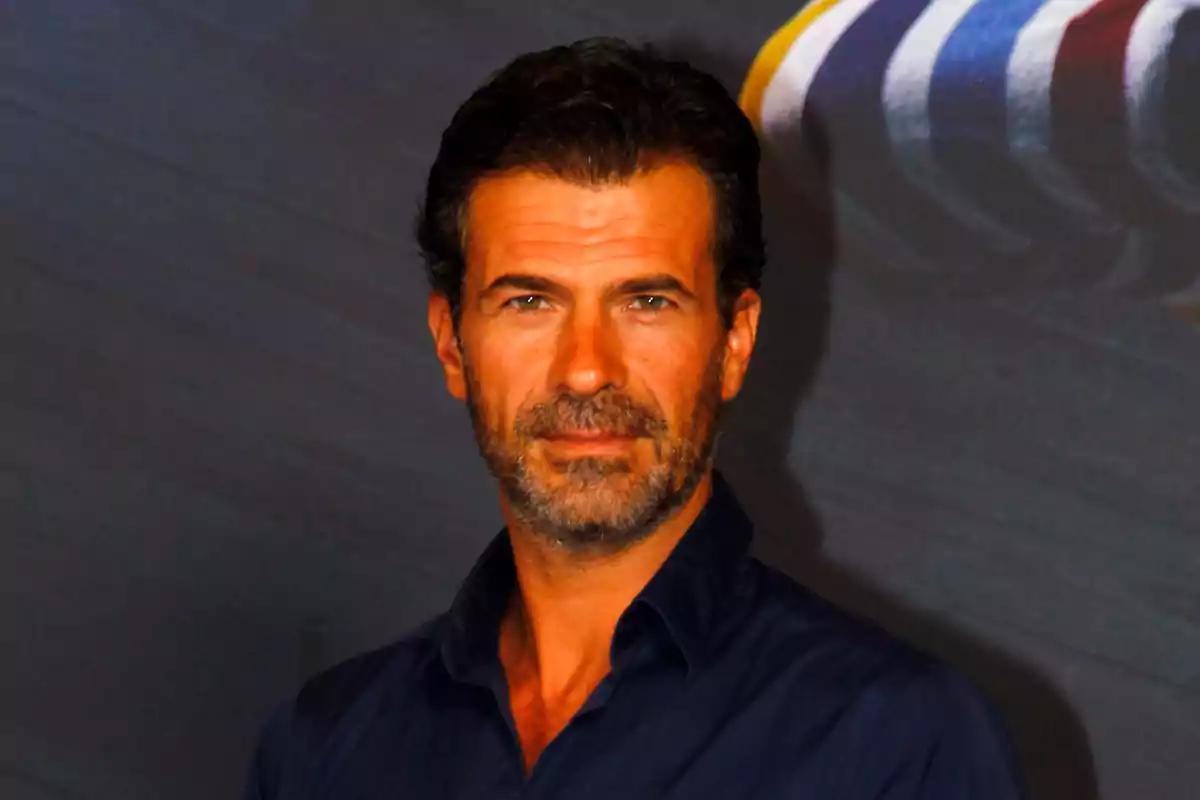 Un hombre con barba y cabello oscuro viste una camisa azul y mira hacia la cámara con una expresión seria.