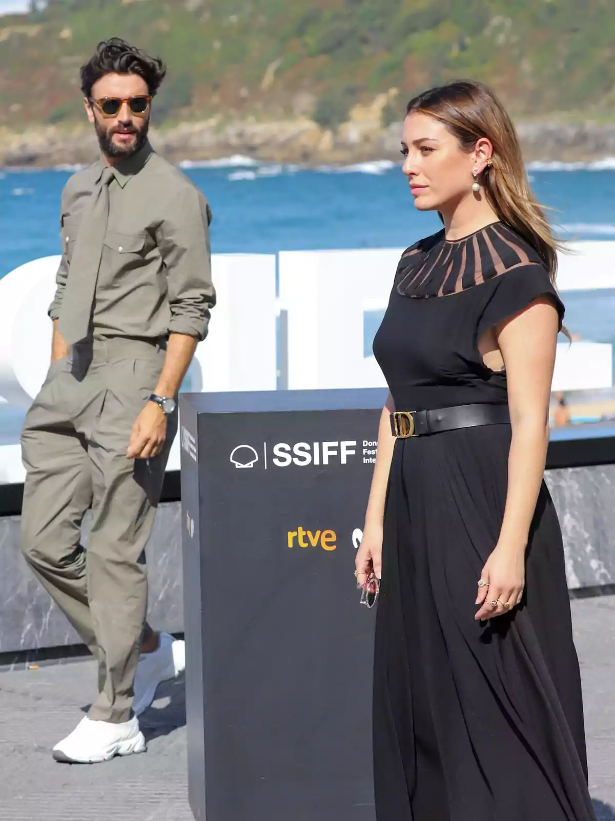 Dos personas posan al aire libre en un evento del Festival Internacional de Cine de San Sebastián, con el mar y colinas verdes de fondo.