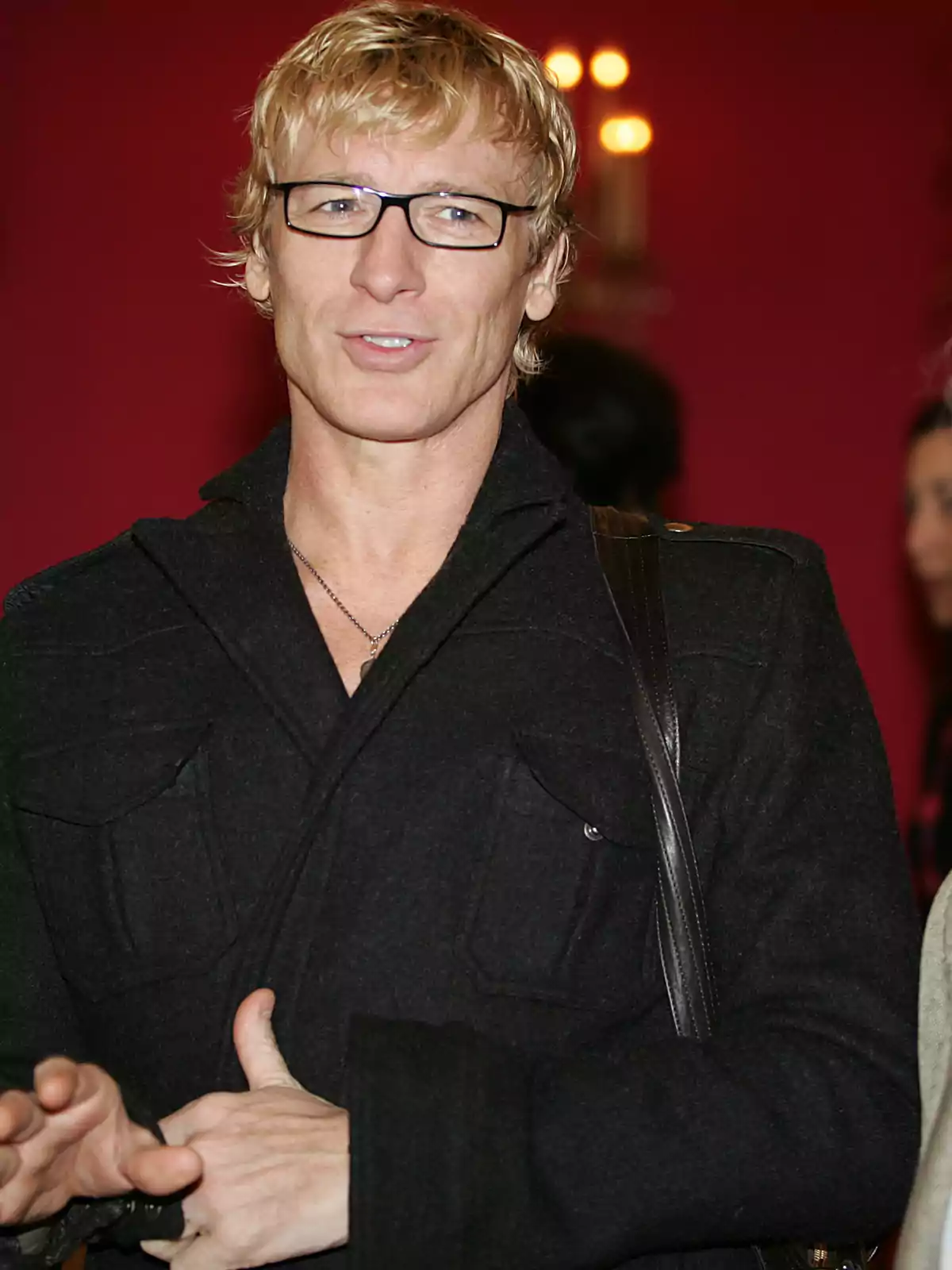 Hombre rubio con gafas y abrigo negro posando frente a un fondo rojo Hombre rubio con gafas y abrigo negro posando frente a un fondo rojo