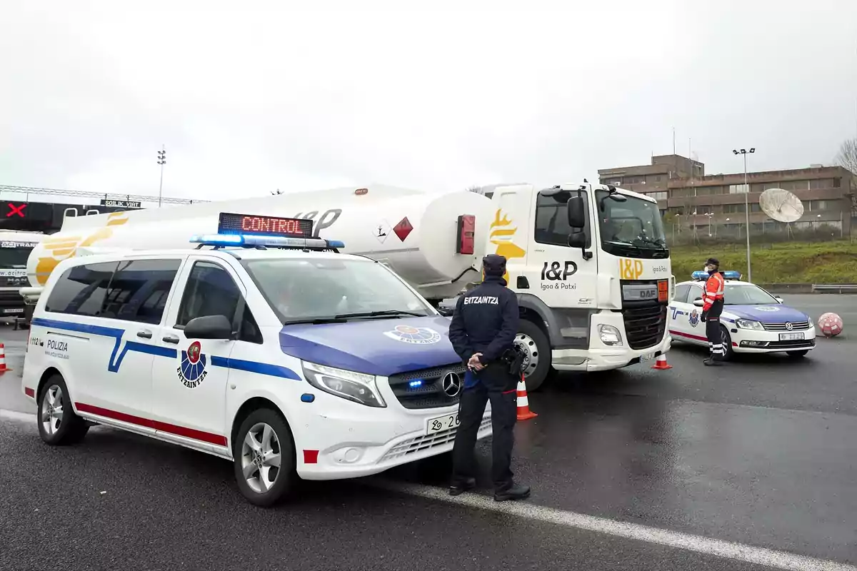 Control policial de carretera con agentes de la Ertzaintza revisando un camión cisterna detenido junto a vehículos patrulla en un día nublado Control policial de carretera con agentes de la Ertzaintza revisando un camión cisterna detenido junto a vehículos patrulla en un día nublado