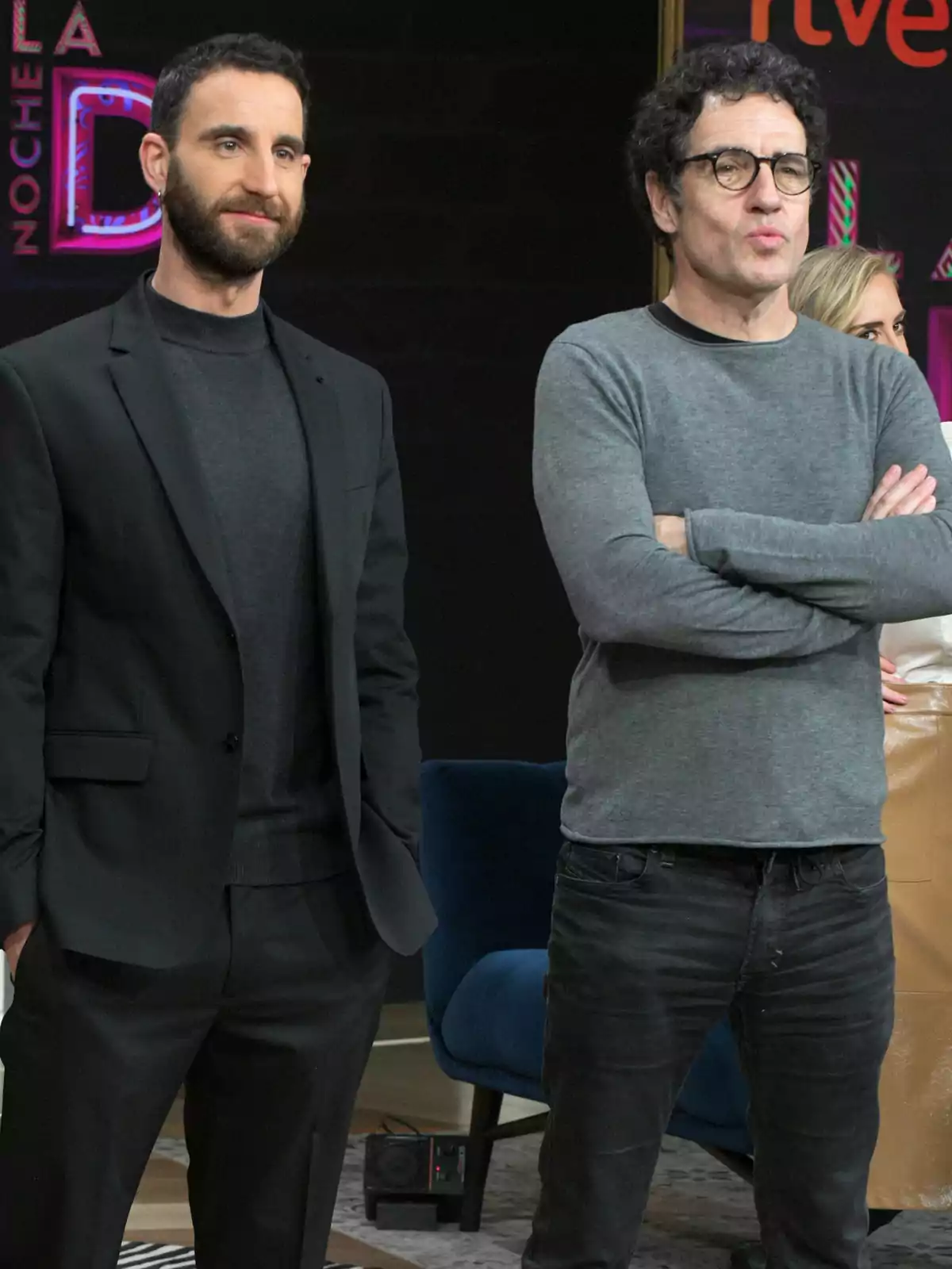 Dos hombres posan juntos en un set de televisión uno lleva traje negro y el otro viste ropa casual con suéter gris y gafas Dos hombres posan juntos en un set de televisión uno lleva traje negro y el otro viste ropa casual con suéter gris y gafas