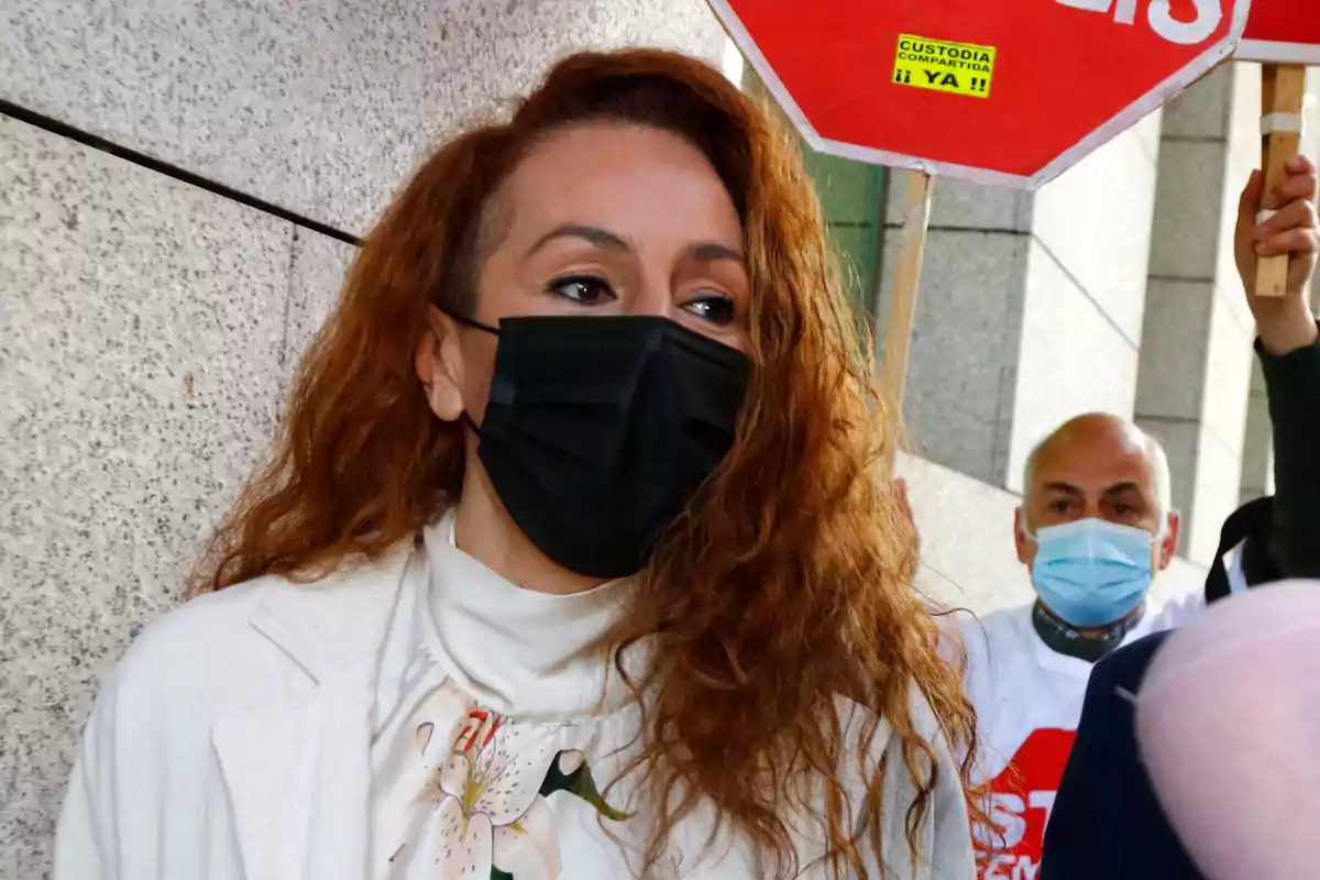 Una persona con mascarilla negra y cabello rizado está en un entorno urbano con otras personas en el fondo que también llevan mascarillas.