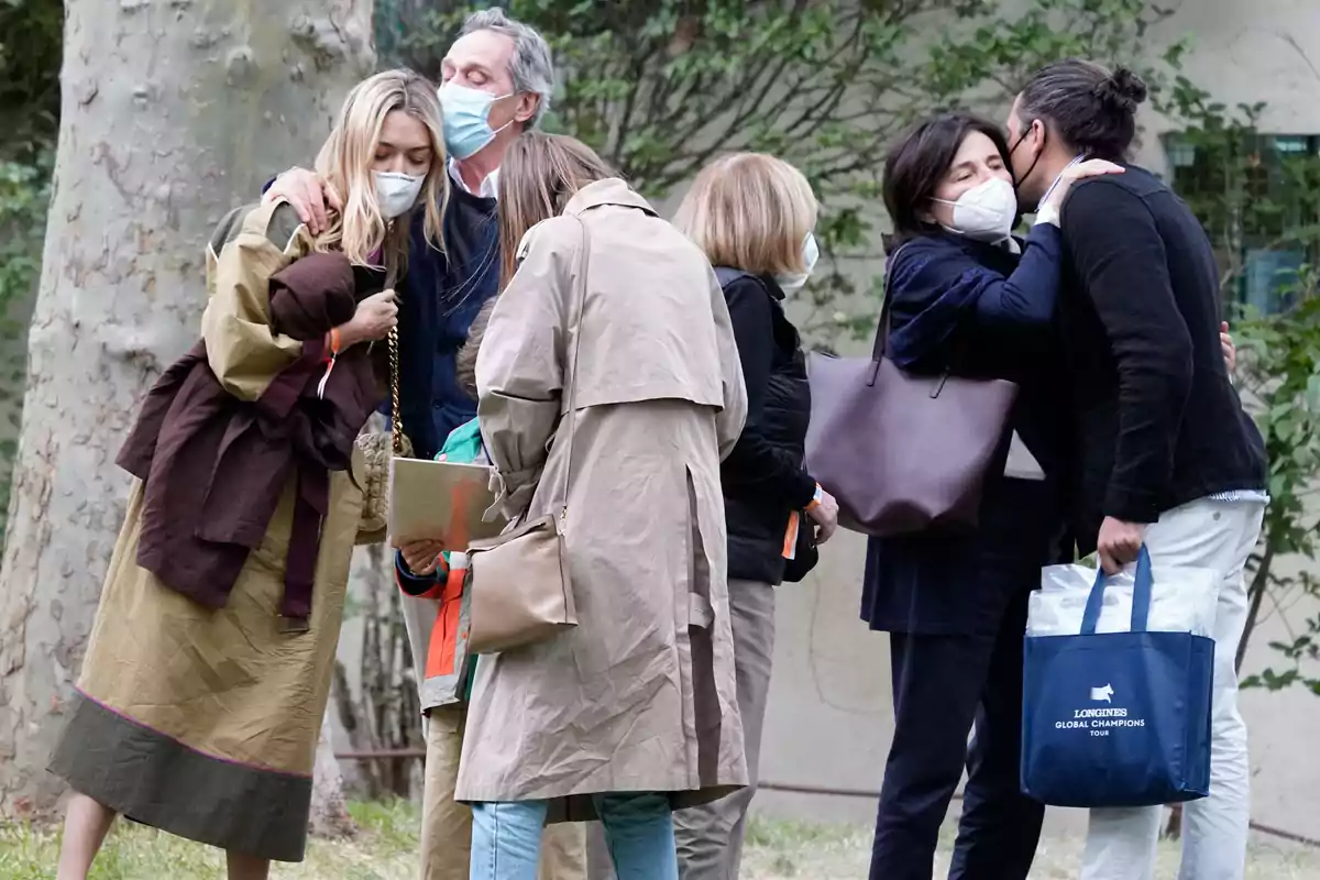 A group of people wearing masks greet each other and talk outdoors in a natural setting; some hug while others look at an object in a woman's hands. A group of people wearing masks greet each other and talk outdoors in a natural setting; some hug while others look at an object in a woman's hands.