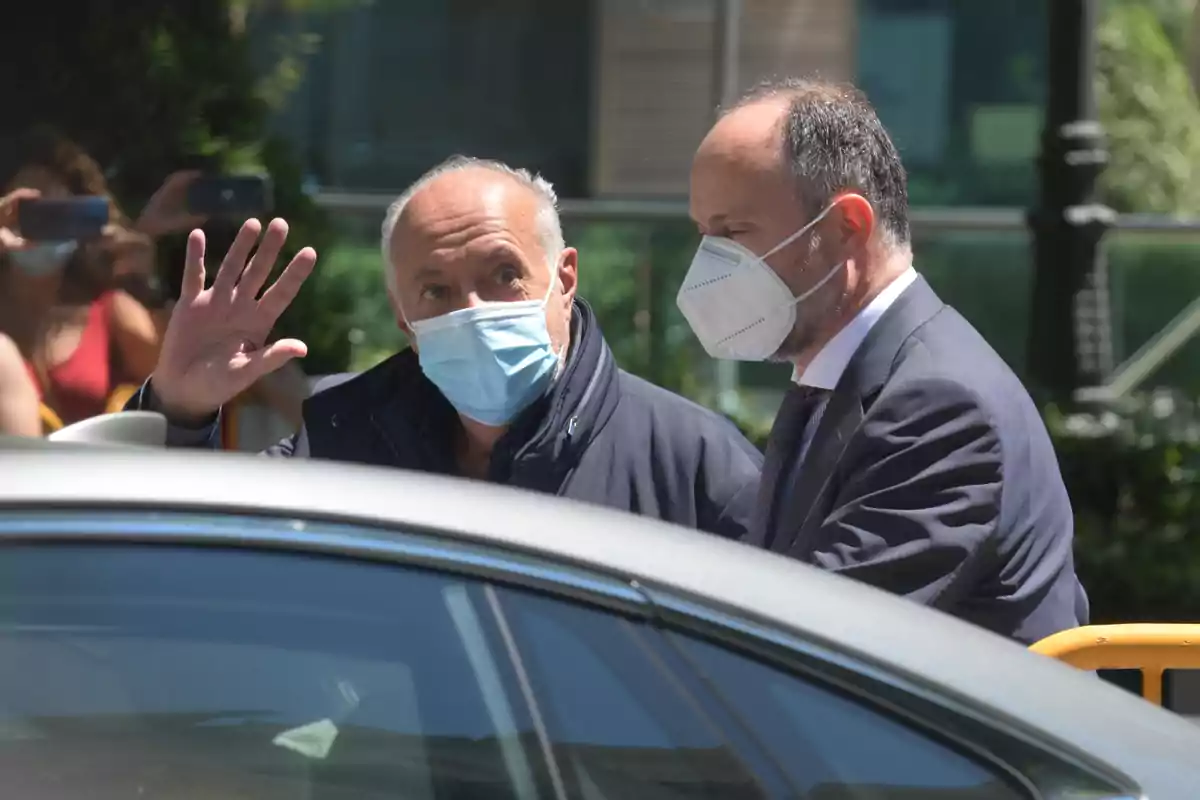 Dos hombres con mascarillas están junto a un coche mientras uno de ellos saluda con la mano.