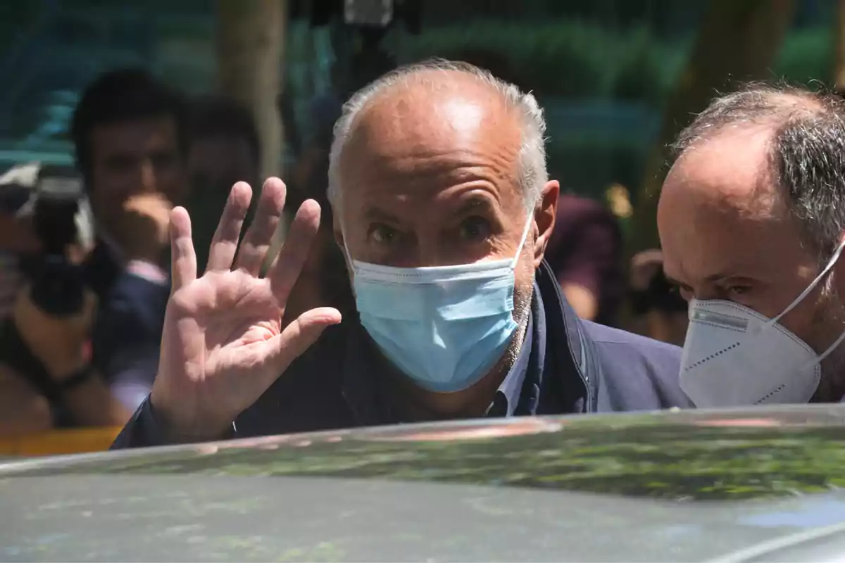 Un hombre con mascarilla azul saluda con la mano mientras está rodeado de personas.