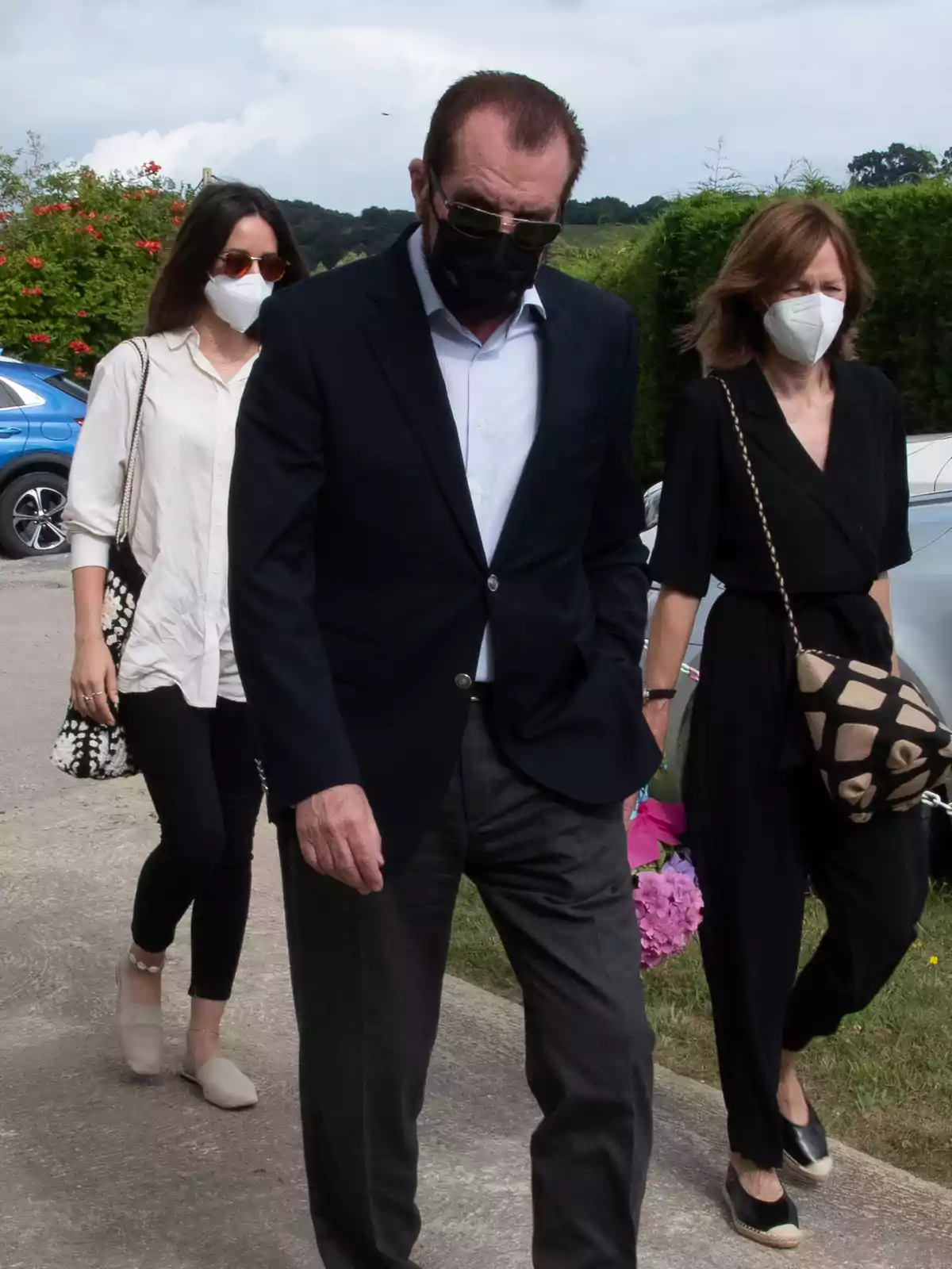 Tres personas con mascarillas caminan al aire libre, una de ellas lleva flores. Tres personas con mascarillas caminan al aire libre, una de ellas lleva flores.