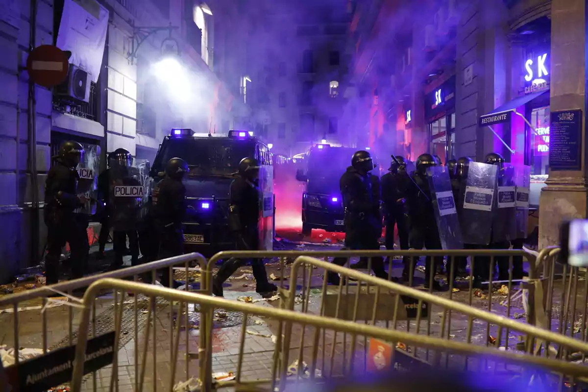 Policías antidisturbios con equipo de protección y escudos se encuentran en una calle llena de escombros y humo, con vehículos policiales y luces azules de emergencia iluminando la escena.