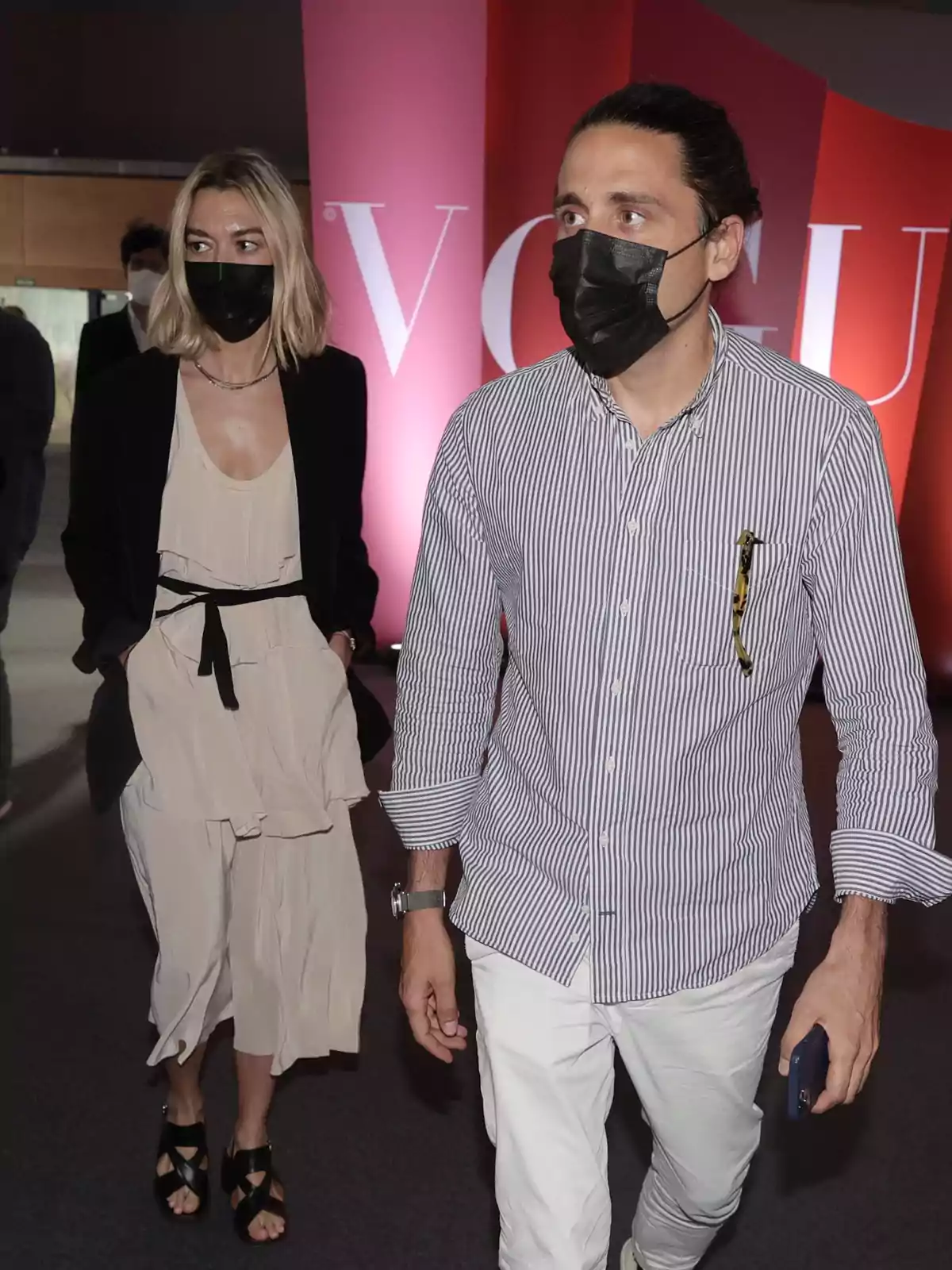 Dos personas con mascarillas caminan frente a un fondo con el logo de una revista. Dos personas con mascarillas caminan frente a un fondo con el logo de una revista.