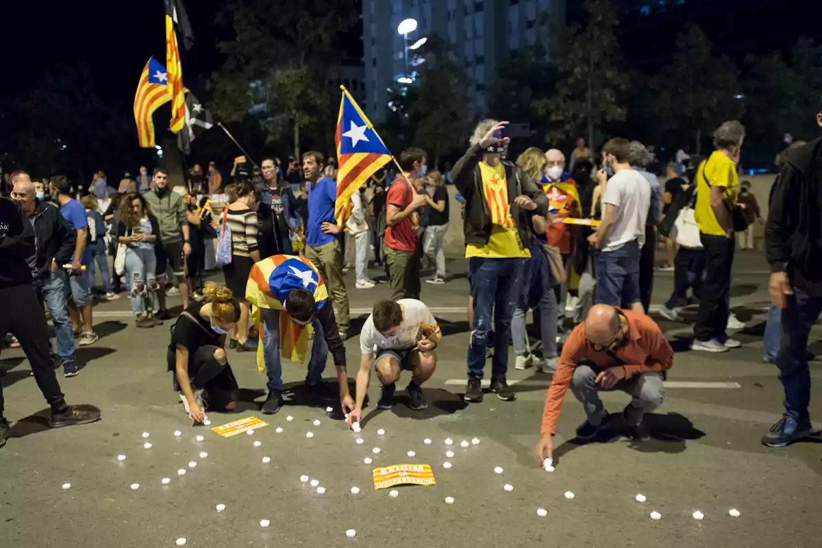 Personas participando en una manifestación nocturna con banderas y velas en el suelo.