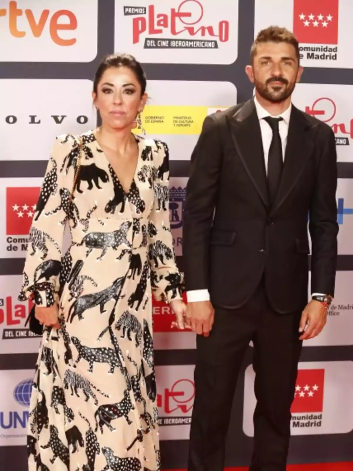 Una pareja posando en la alfombra roja de los Premios Platino del Cine Iberoamericano, ella lleva un vestido largo con estampado de animales y él un traje negro con corbata.