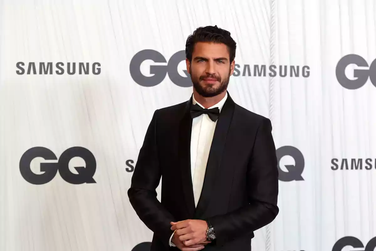 Hombre con esmoquin negro posando en un evento de GQ con logotipos de Samsung y GQ en el fondo.