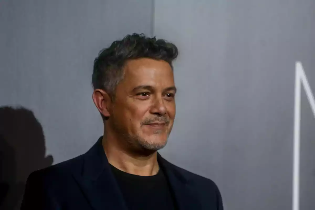 Hombre de cabello corto y canoso con barba ligera vistiendo saco oscuro y camiseta negra posando frente a un fondo gris Hombre de cabello corto y canoso con barba ligera vistiendo saco oscuro y camiseta negra posando frente a un fondo gris