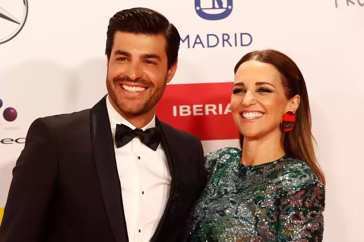 Pareja sonriente posando en una alfombra roja con fondo de logotipos y ambos vestidos de gala Pareja sonriente posando en una alfombra roja con fondo de logotipos y ambos vestidos de gala