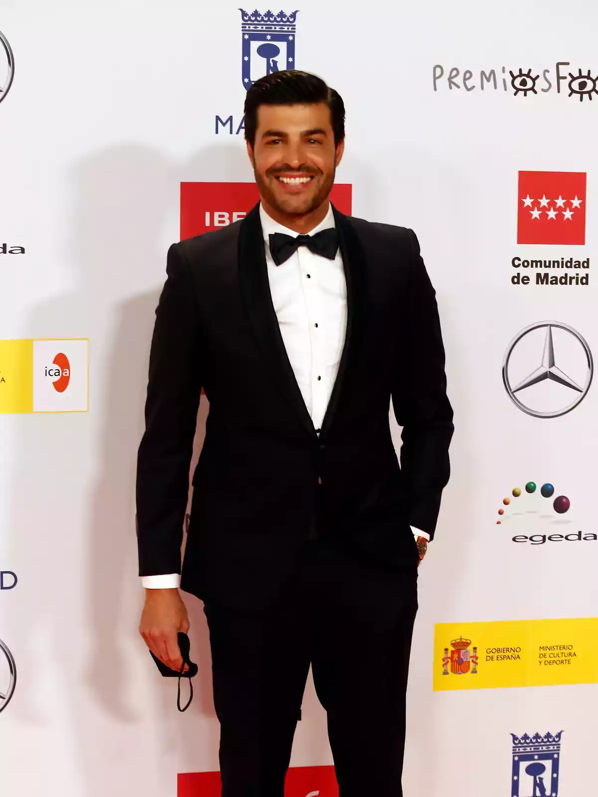 Hombre con esmoquin negro y pajarita posando sonriente en una alfombra roja frente a un fondo con logotipos de patrocinadores y entidades oficiales Hombre con esmoquin negro y pajarita posando sonriente en una alfombra roja frente a un fondo con logotipos de patrocinadores y entidades oficiales
