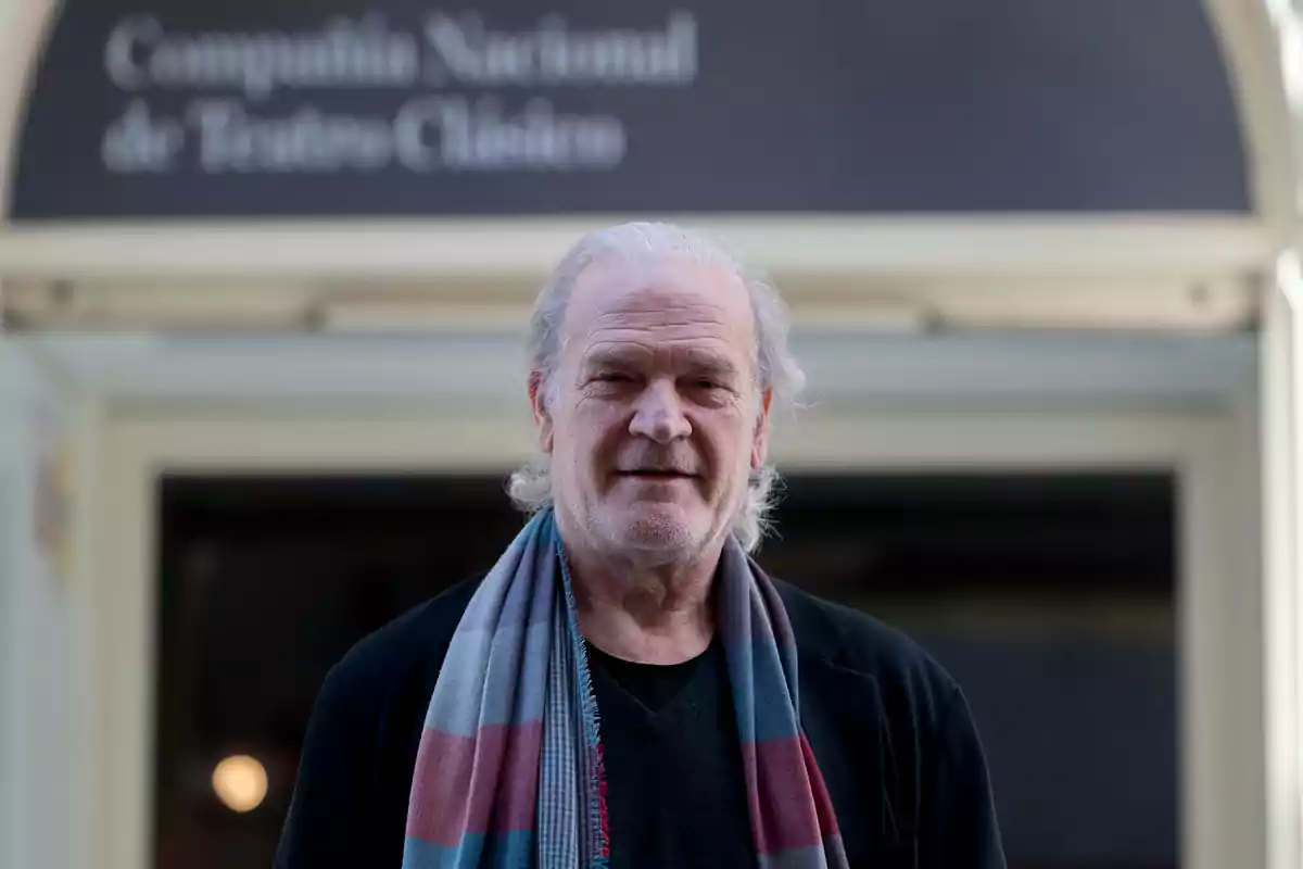 Un hombre de cabello canoso y bufanda está de pie frente a un edificio con un cartel que dice "Compañía Nacional de Teatro Clásico".