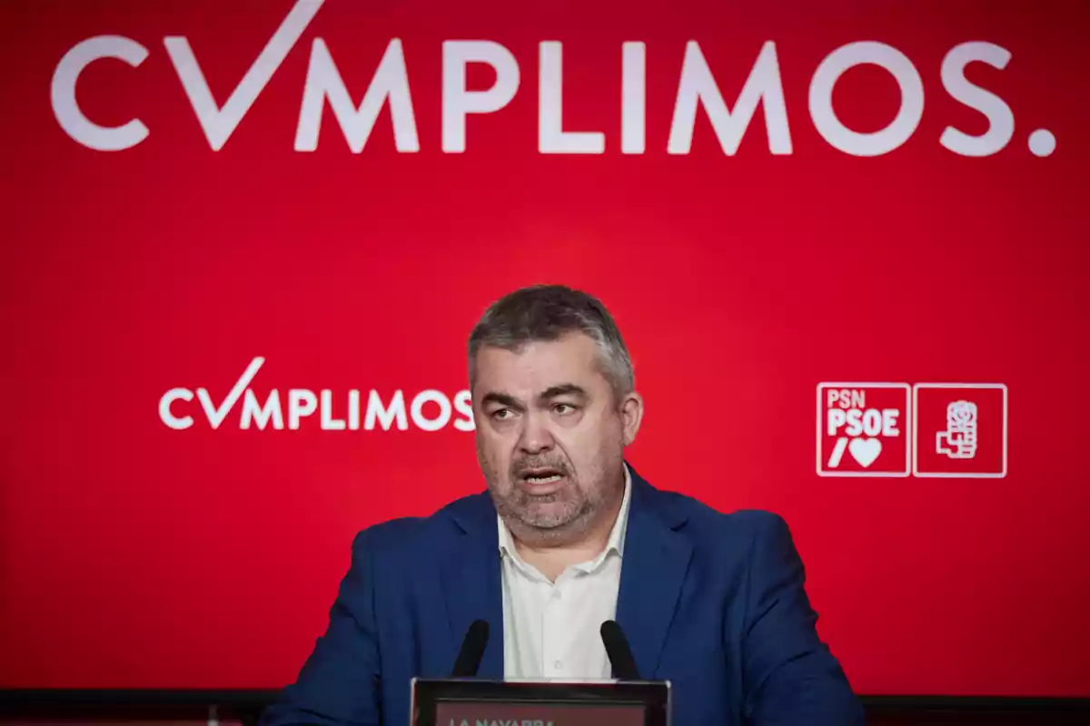 Hombre de cabello canoso y barba hablando en una rueda de prensa frente a un fondo rojo con el texto cumplimos y logotipos del partido PSN PSOE Hombre de cabello canoso y barba hablando en una rueda de prensa frente a un fondo rojo con el texto cumplimos y logotipos del partido PSN PSOE