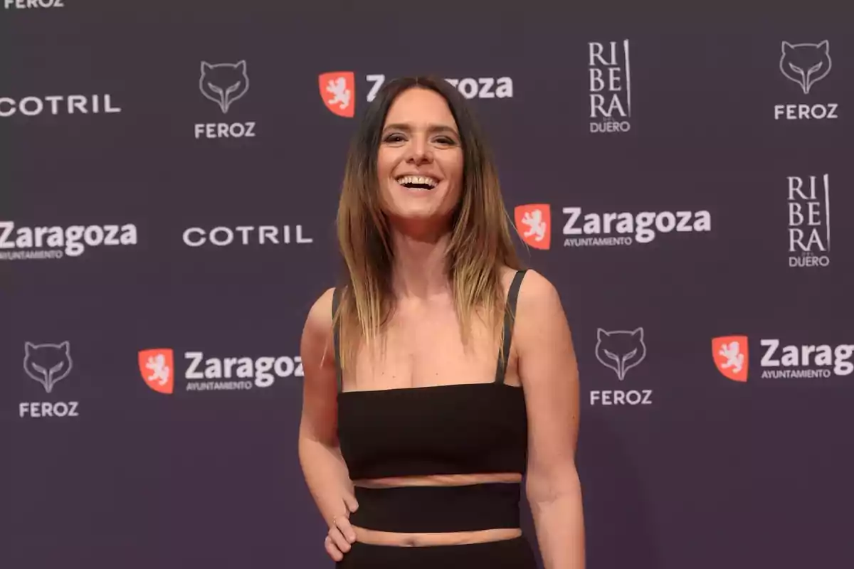 Una persona sonriendo en un evento con un fondo que muestra varios logotipos y nombres de marcas. Una persona sonriendo en un evento con un fondo que muestra varios logotipos y nombres de marcas.