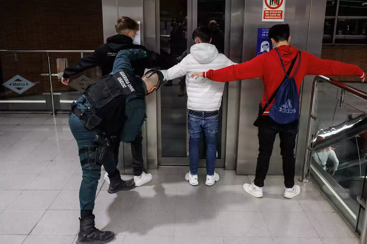 Un agente de la Guardia Civil revisa a tres jóvenes con los brazos extendidos frente a un ascensor en una estación de metro Un agente de la Guardia Civil revisa a tres jóvenes con los brazos extendidos frente a un ascensor en una estación de metro