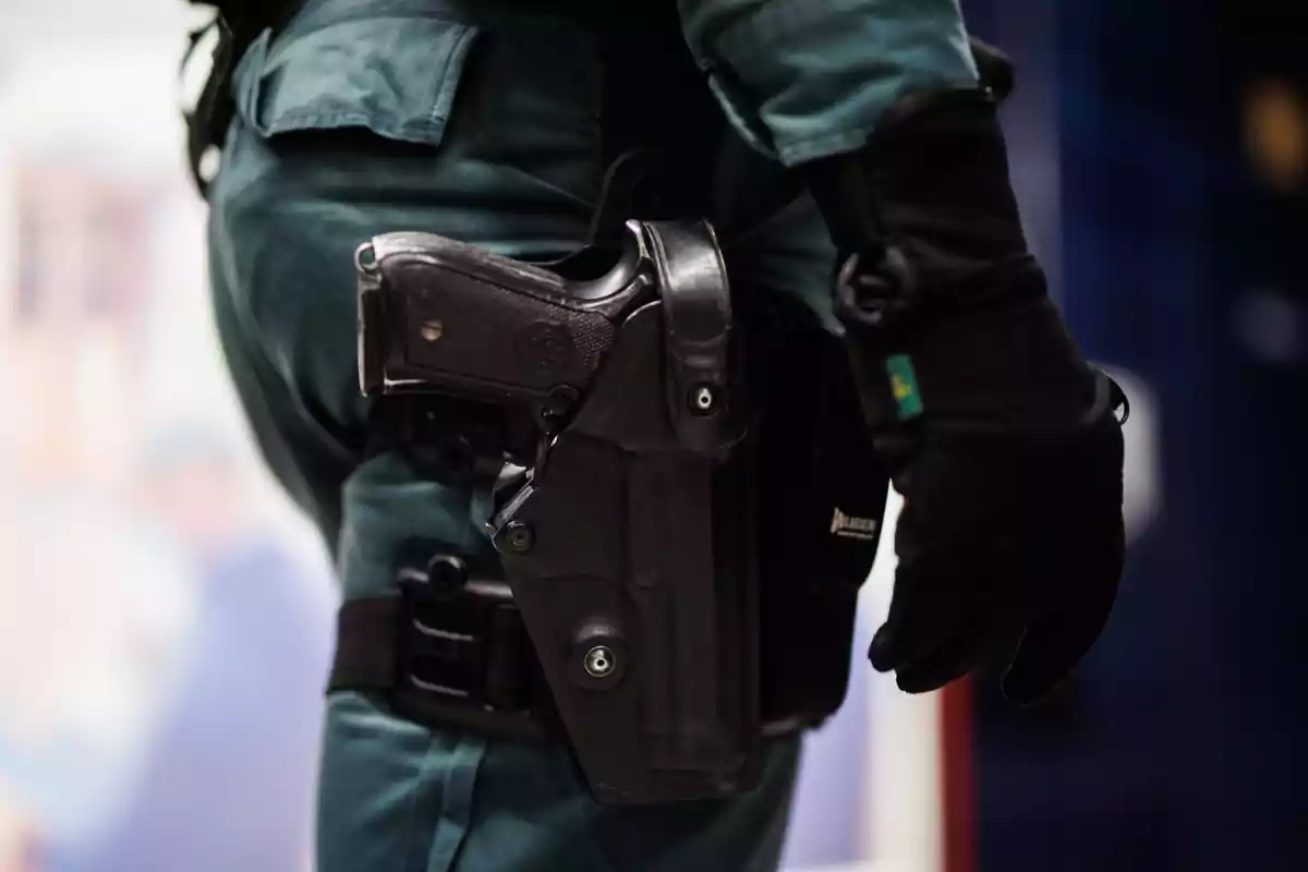 Persona con uniforme verde y guantes negros lleva una pistola enfundada en la cintura Persona con uniforme verde y guantes negros lleva una pistola enfundada en la cintura