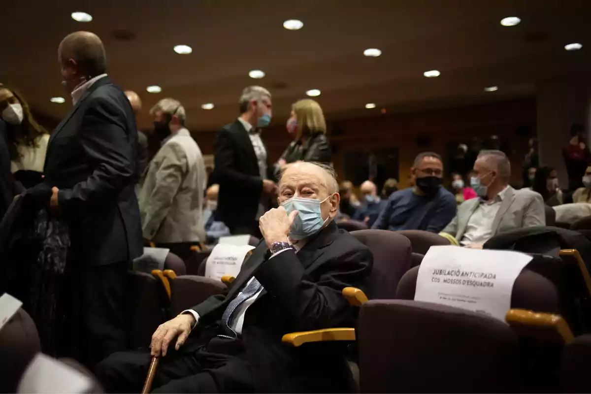 Una persona mayor con mascarilla está sentada en un auditorio rodeado de otras personas que también llevan mascarillas.