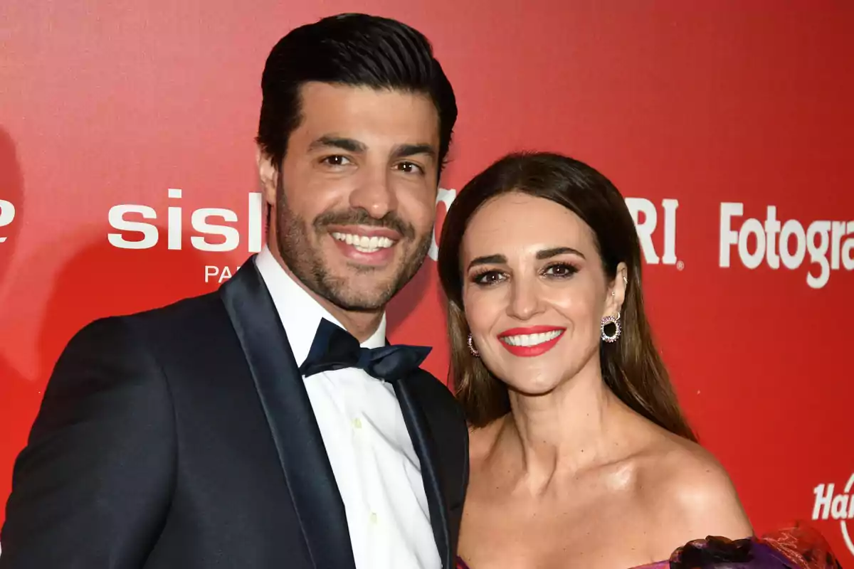 Pareja sonriendo elegantemente vestida en un evento con fondo rojo y logotipos blancos Pareja sonriendo elegantemente vestida en un evento con fondo rojo y logotipos blancos