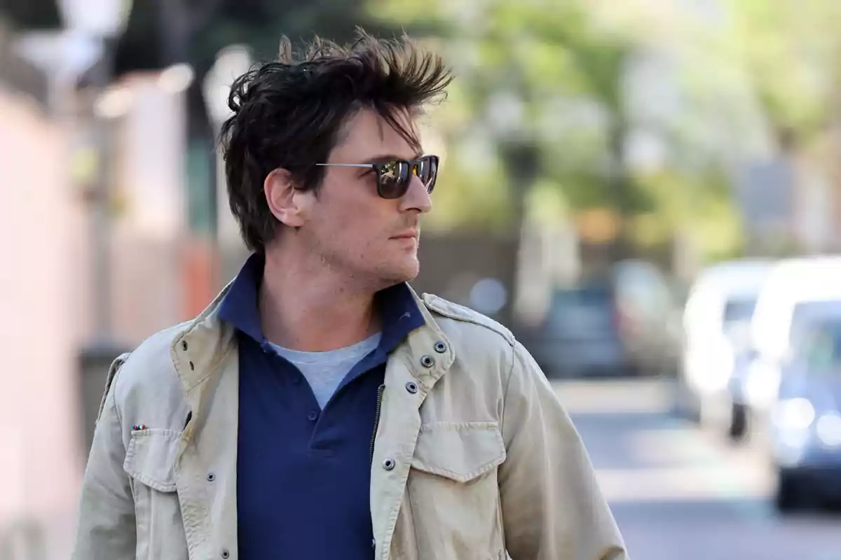 Un hombre con gafas de sol y chaqueta beige camina por una calle urbana.