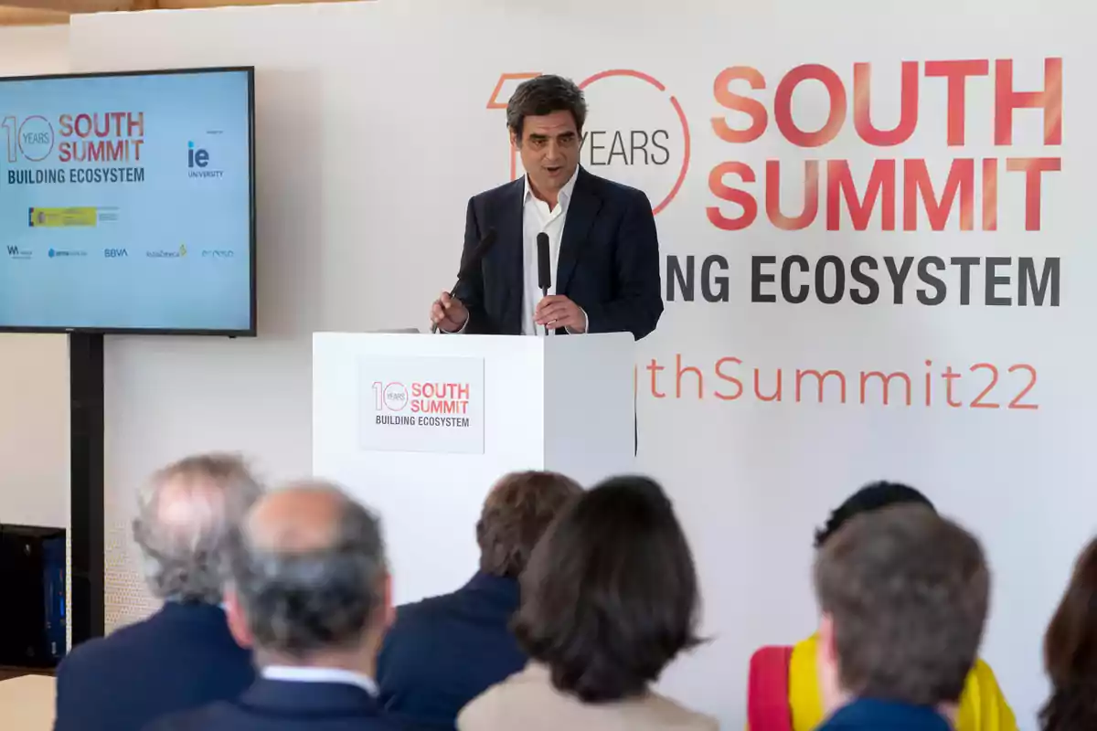 Un hombre hablando en un podio durante el South Summit 2022 con una pantalla y un cartel del evento en el fondo.