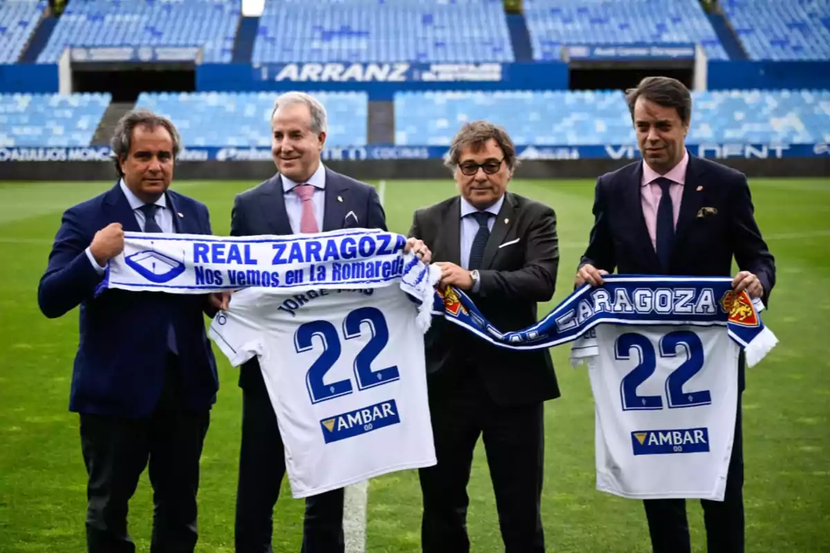 Cuatro hombres en traje sostienen bufandas y camisetas del Real Zaragoza en un estadio de fútbol vacío.