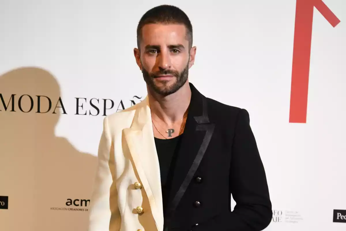 Un hombre con barba y cabello corto, vestido con un traje de dos colores, posa frente a un fondo blanco con texto.