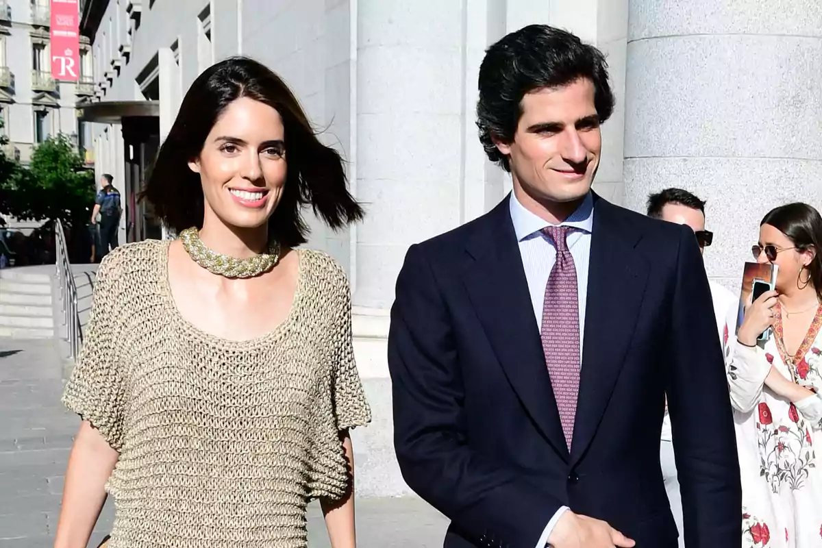 Una mujer y un hombre caminando juntos al aire libre, ella lleva un suéter beige y un collar dorado, mientras que él viste un traje oscuro con corbata.