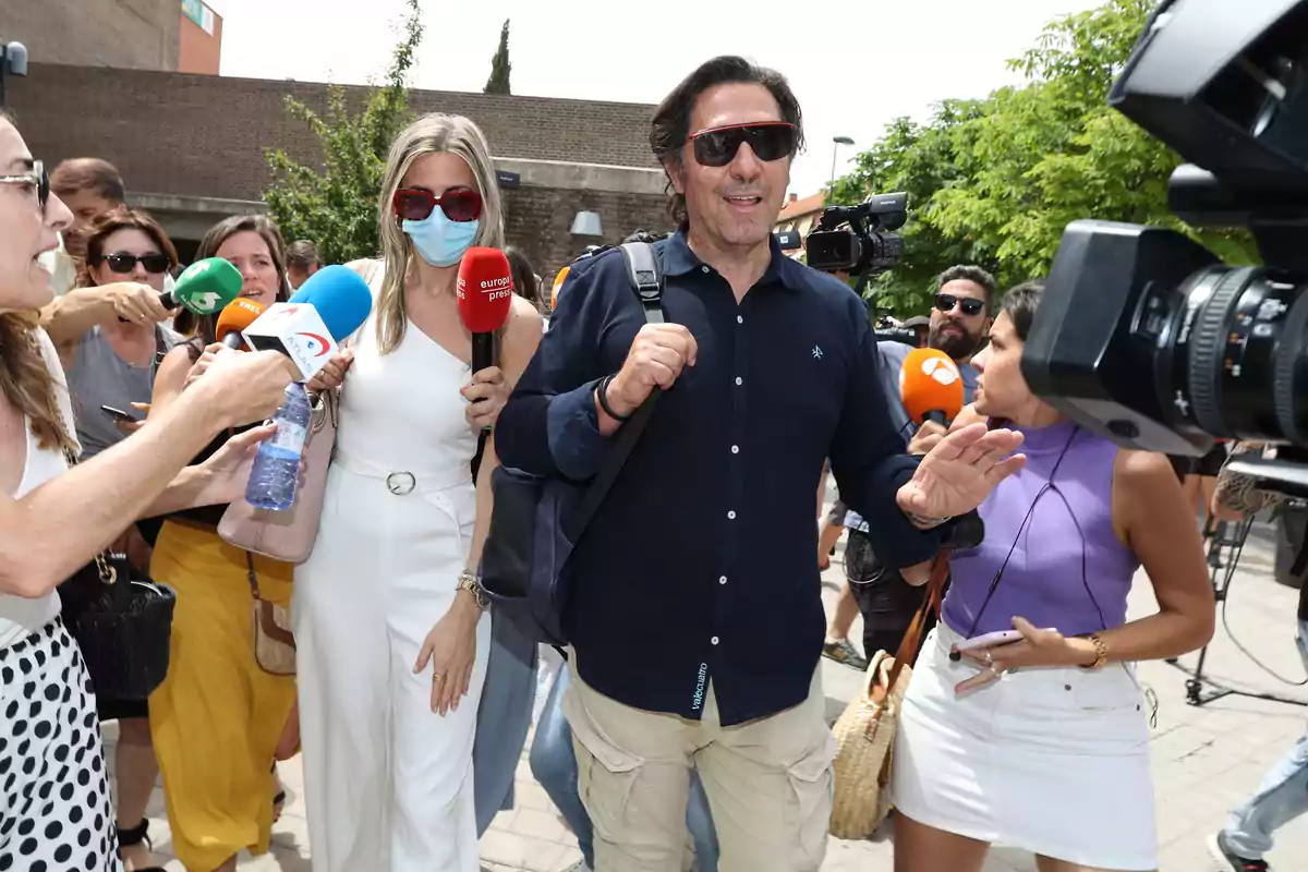 Un hombre con gafas de sol y una mochila es rodeado por periodistas y camarógrafos mientras camina al aire libre.