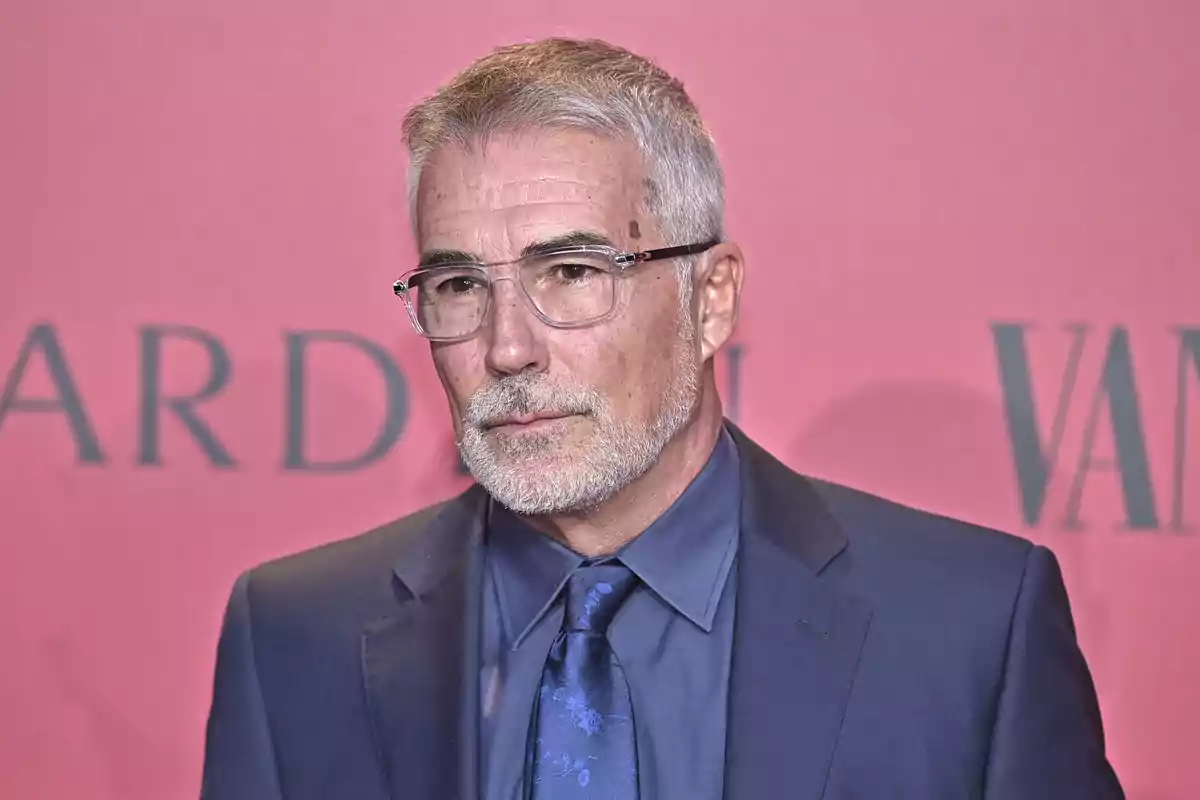 Hombre con barba y gafas vistiendo un traje azul frente a un fondo rojo.