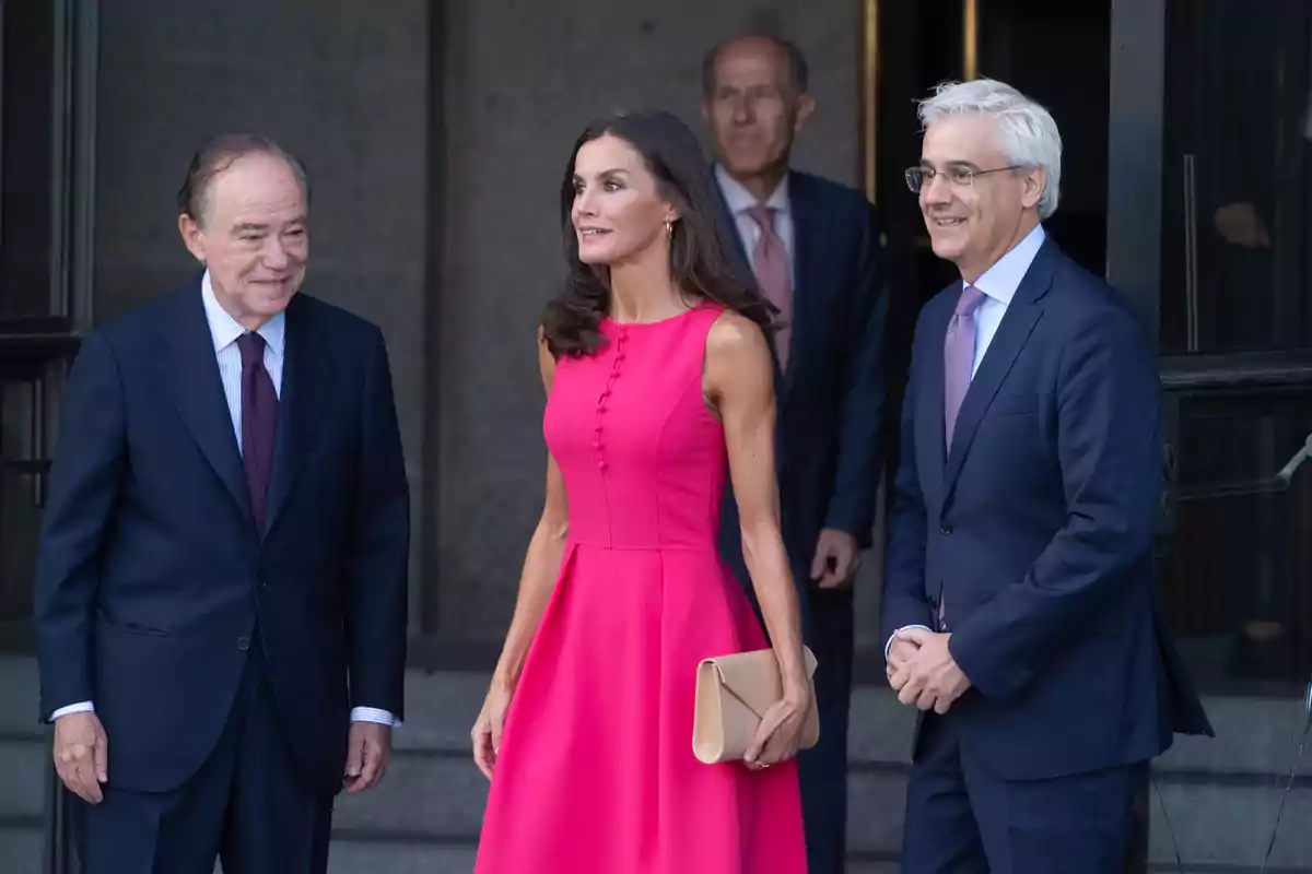 Un grupo de personas vestidas formalmente, una mujer con un vestido rosa y dos hombres con trajes oscuros, están de pie frente a un edificio.