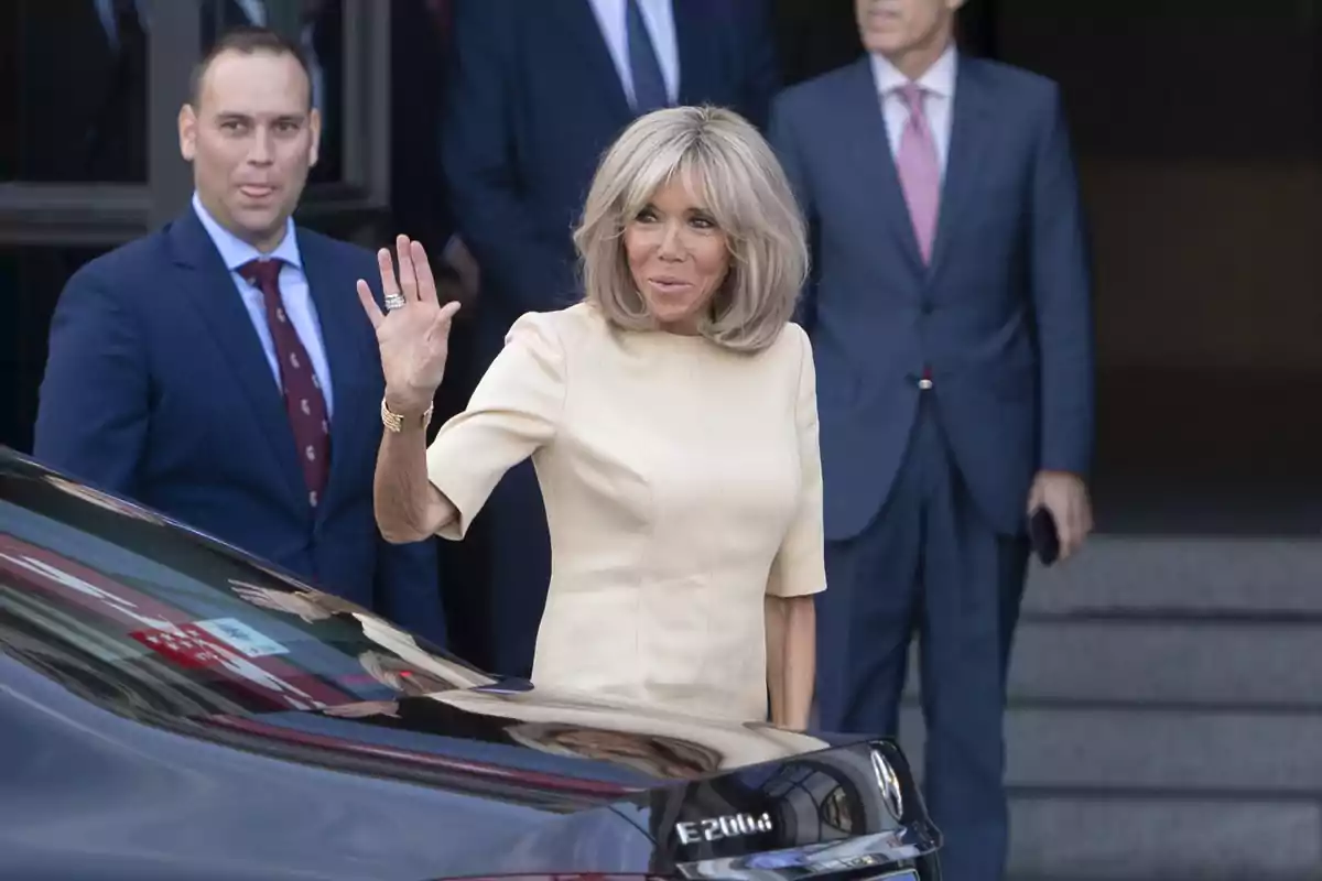 Mujer rubia con vestido claro saluda con la mano mientras se encuentra junto a un automóvil negro y varias personas con traje al fondo
