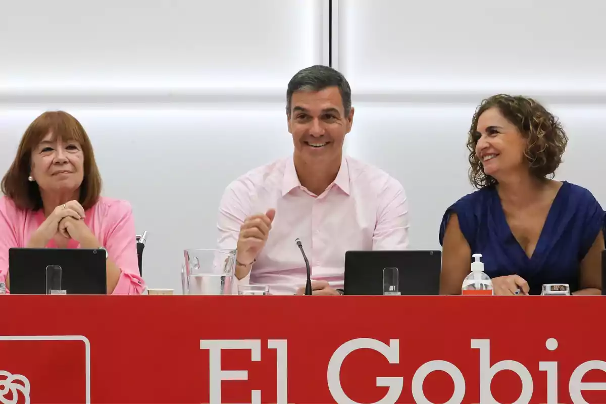 Tres personas sentadas en una mesa roja durante una reunión, dos mujeres a los lados y un hombre en el centro, todos sonrientes y con dispositivos electrónicos frente a ellos. Tres personas sentadas en una mesa roja durante una reunión, dos mujeres a los lados y un hombre en el centro, todos sonrientes y con dispositivos electrónicos frente a ellos.