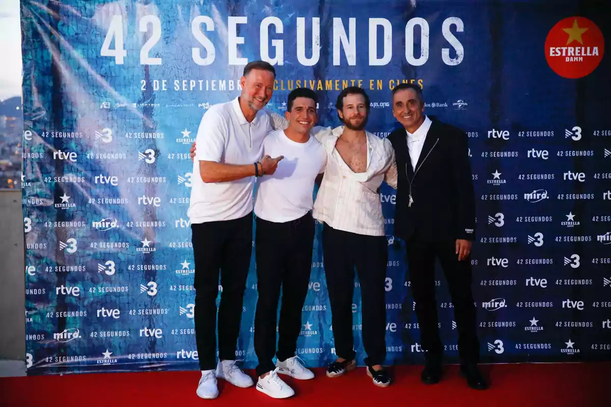 Cuatro hombres posan juntos en una alfombra roja frente a un fondo promocional de la película 