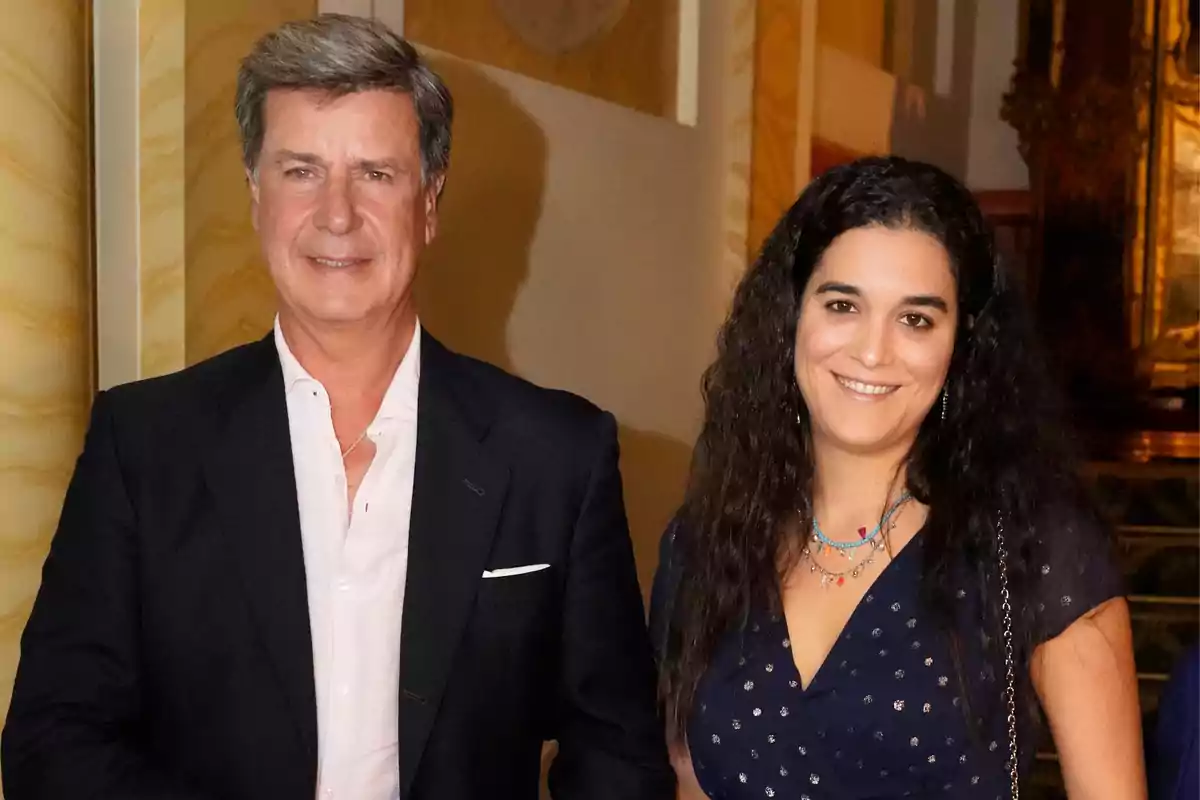 Un hombre de cabello canoso y una mujer de cabello largo y oscuro sonríen juntos en un elegante interior con paredes decoradas.